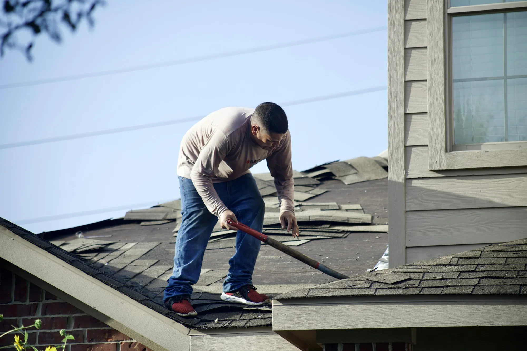 Worker prying up old shingles on a damaged house roof